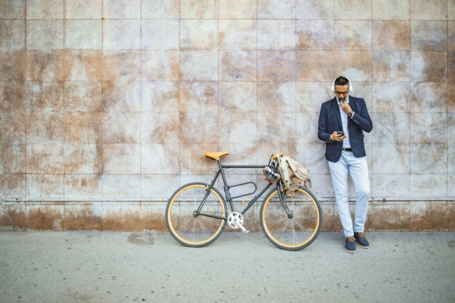 man leaning against a wall next to a bicycle wearing headphones looking at his mobile phone