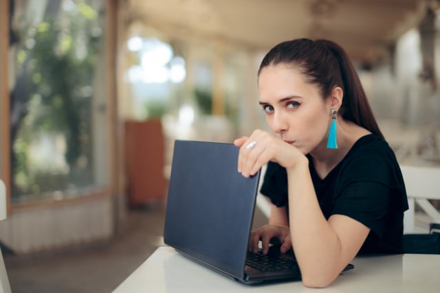 photo of a woman sitting at a table with a laptop, shielding her screen from potential snoopers