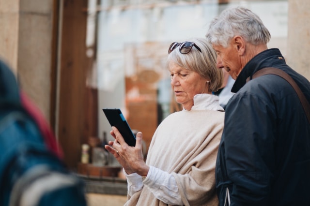 photo of an older woman and older man in the street, the woman holds a mobile phone and is looking at it, the man is looking at it over the woman's shoulder