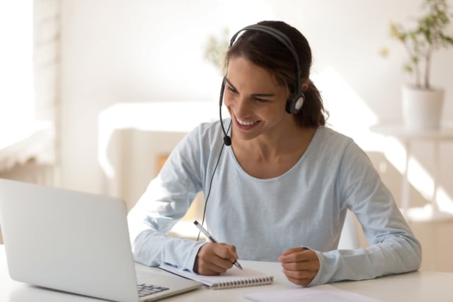 woman working at a table from home, wearing a headset and writing on a notepad while smiling at her laptop