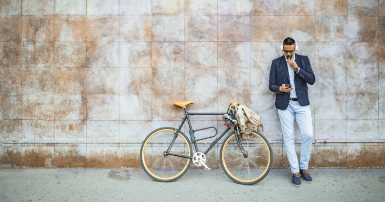 man leaning against a wall next to a bicycle wearing headphones looking at his mobile phone