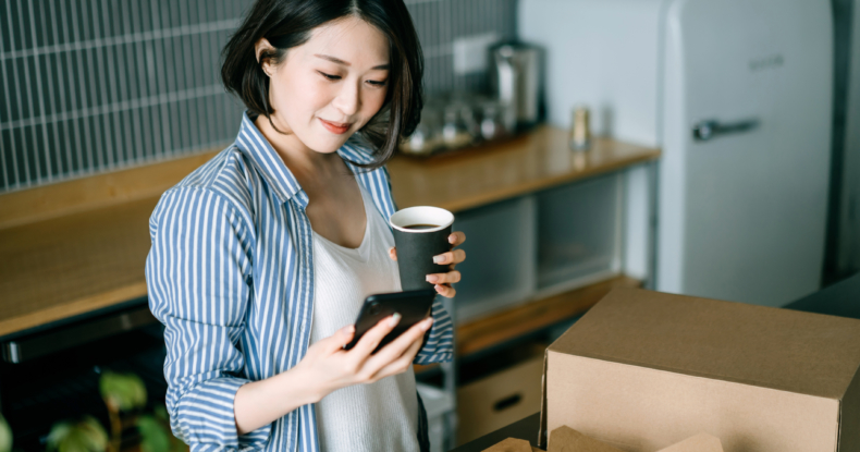 Beautiful smiling young Asian woman standing by the kitchen counter, using smartphone while enjoying assorted home delivery takeaway food, coffee and fresh green salad at home. Eating at home concept-273